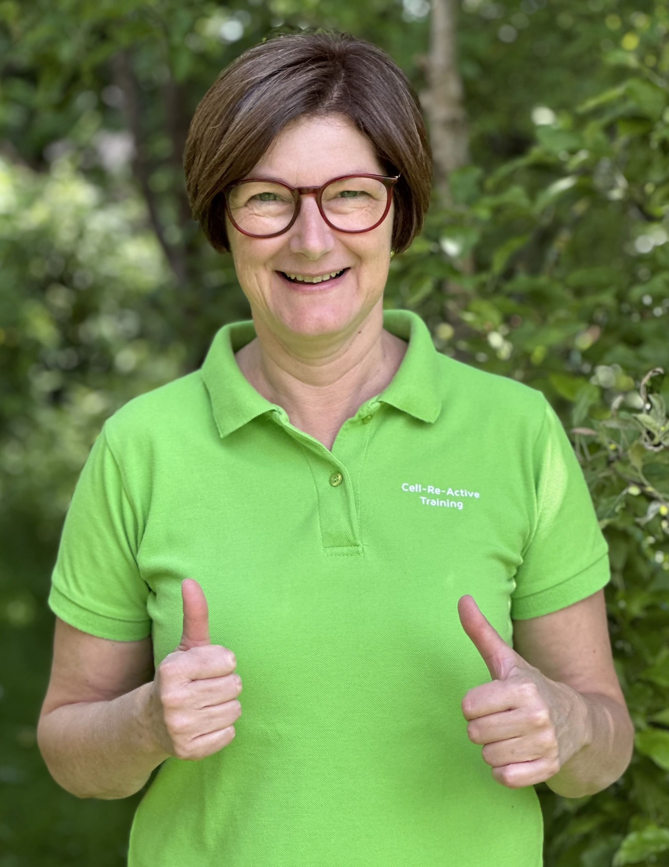Olga Lebenbauer, cell-re-active Trainerin & FNL Kräuterexpertin Olga Lebenbauer mit grünem T-Shirt, lächelt und zeigt beide Daumen hoch – Ausdruck von Freude und Motivation als Cell-re-active-Trainerin